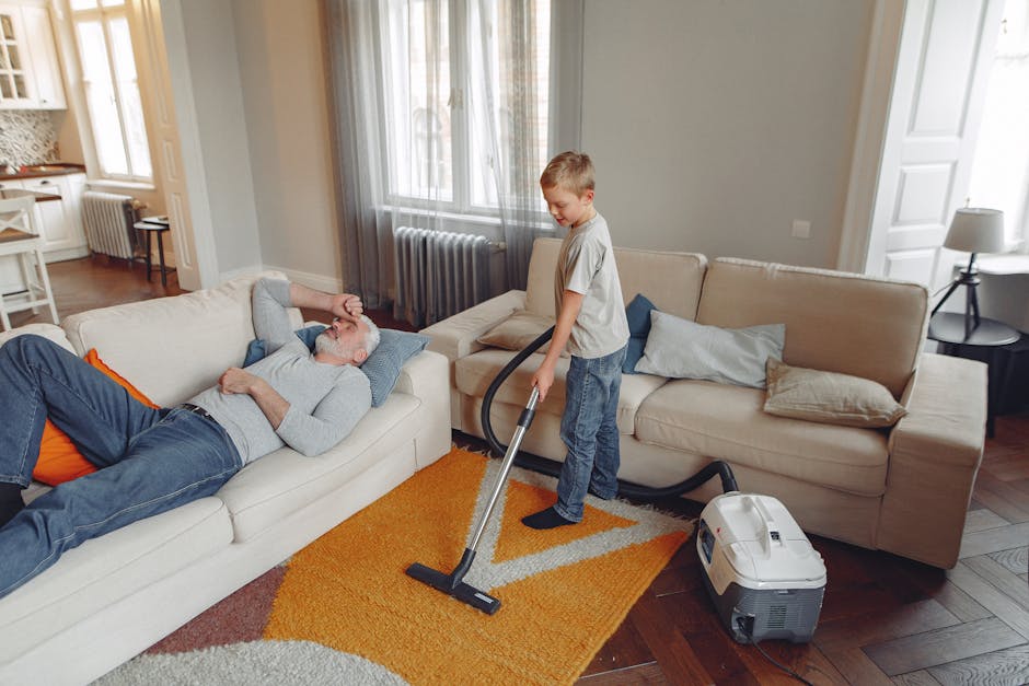 Close-up image of a person vacuuming a patterned, traditional-style carpet using a black upright vacuum cleaner with a wide cleaning head. The carpet features floral motifs in shades of beige, green,