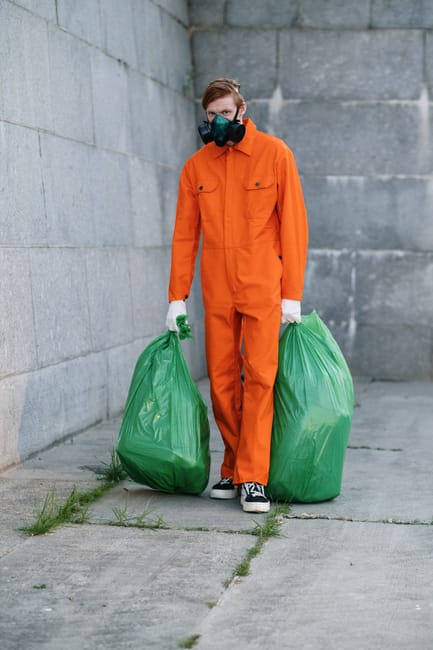 An overflowing public waste bin situated on a paved sidewalk in front of a commercial building, with a variety of discarded rubbish including cardboard boxes, plastic bags, paper, and packaging materi